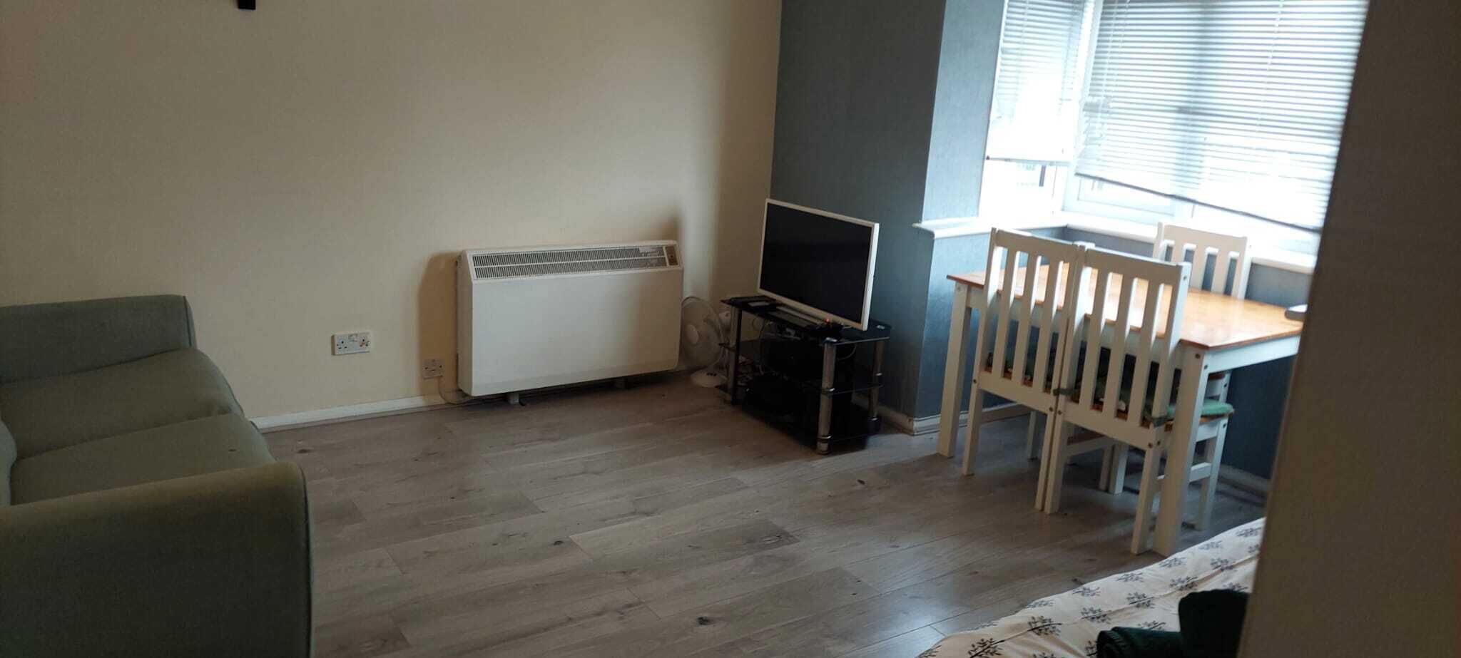 Un salon avec un canapé, un téléviseur posé sur un meuble, un radiateur et une fenêtre avec des stores.