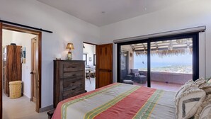Ein Schlafzimmer mit einem großen Bett, einer Holzkommode und Blick auf den Ozean durch eine Schiebetür.