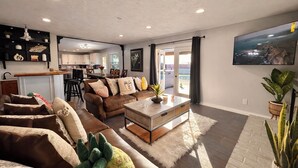 Ein geräumiges Wohnzimmer mit einer großen Couch, einem Couchtisch und einem an der Wand befestigten Fernseher. Im Hintergrund ist eine Küche mit weißen Schränken und einem Essbereich zu sehen.