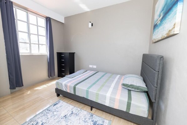 A bedroom with a bed, a dresser, a window with curtains, and a painting on the wall. - Tranquil 2-bedroom apartment with AC in charming Roseau (Roseau)
