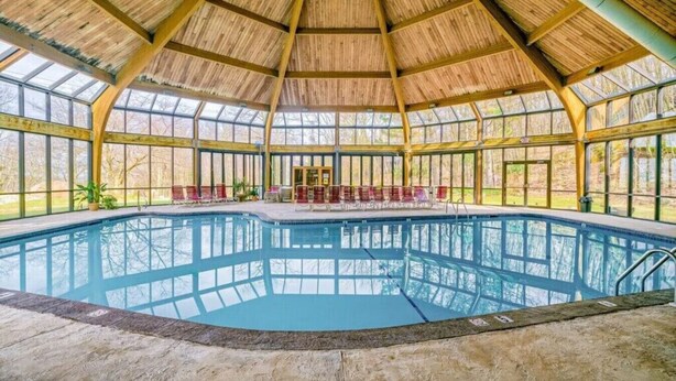 Uma grande piscina interior com cobertura de madeira e paredes de vidro, rodeada por áreas de estar.