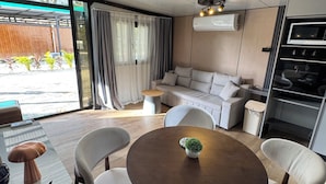 A modern living room with a round wooden table, white cushioned chairs, a sofa, and a small potted plant.