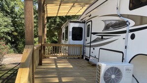 A covered deck with a wooden railing, an air conditioning unit, and a recreational vehicle parked in the background. - RV #1 - delightful 1-bdrm, 3 slide out, covered deck, at Lake Livingston Tx  (Onalaska)