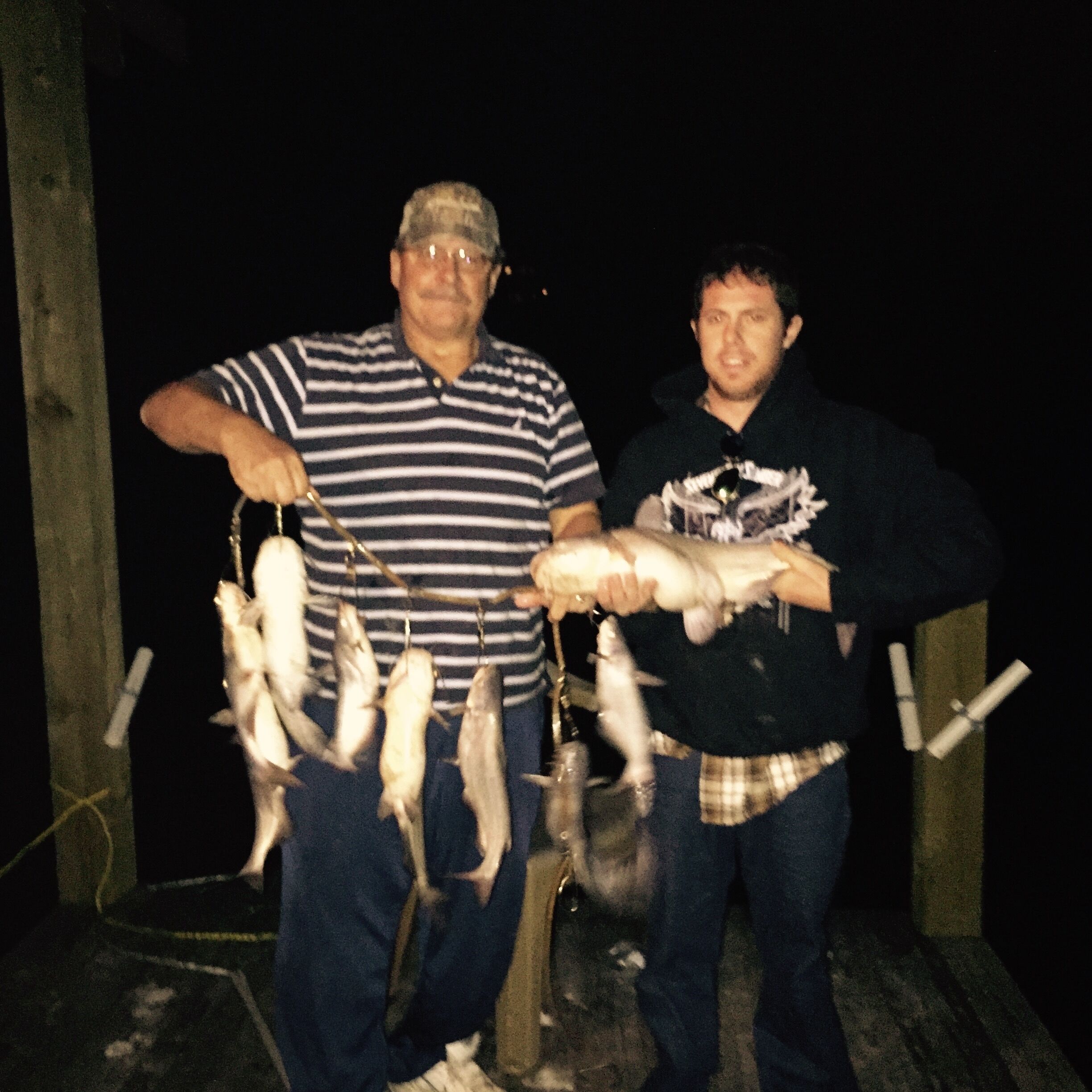 Two individuals holding fish, standing in a dimly lit area with a wooden structure in the background.