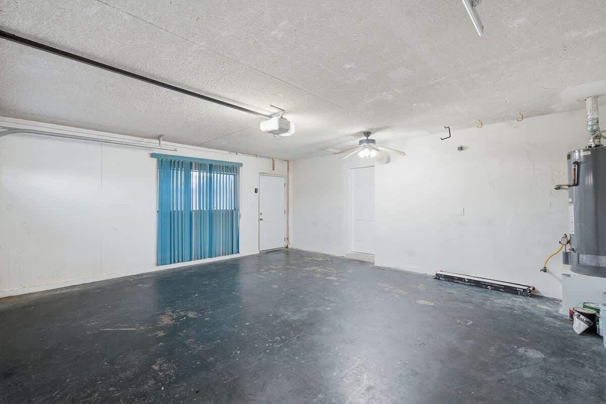Una habitación amplia y vacía con piso negro, paredes blancas, un ventilador de techo y una puerta con cortinas azules.