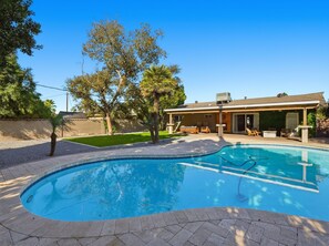 Ein Pool mit gewölbter Kante, eine überdachte Terrasse mit Gartenmöbeln und ein Haus mit geneigtem Dach und großen Fenstern.