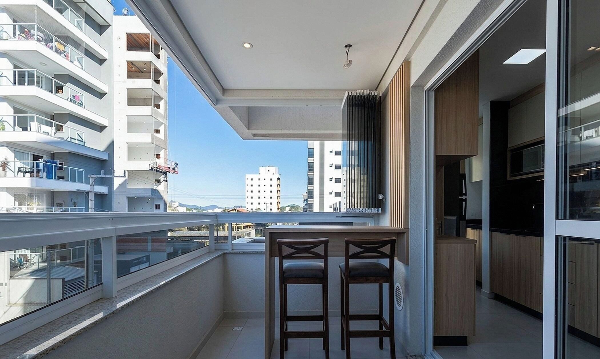 Un balcón con vistas a edificios cercanos, una mesa pequeña con dos sillas y un ventanal corredero que da a una zona de cocina.