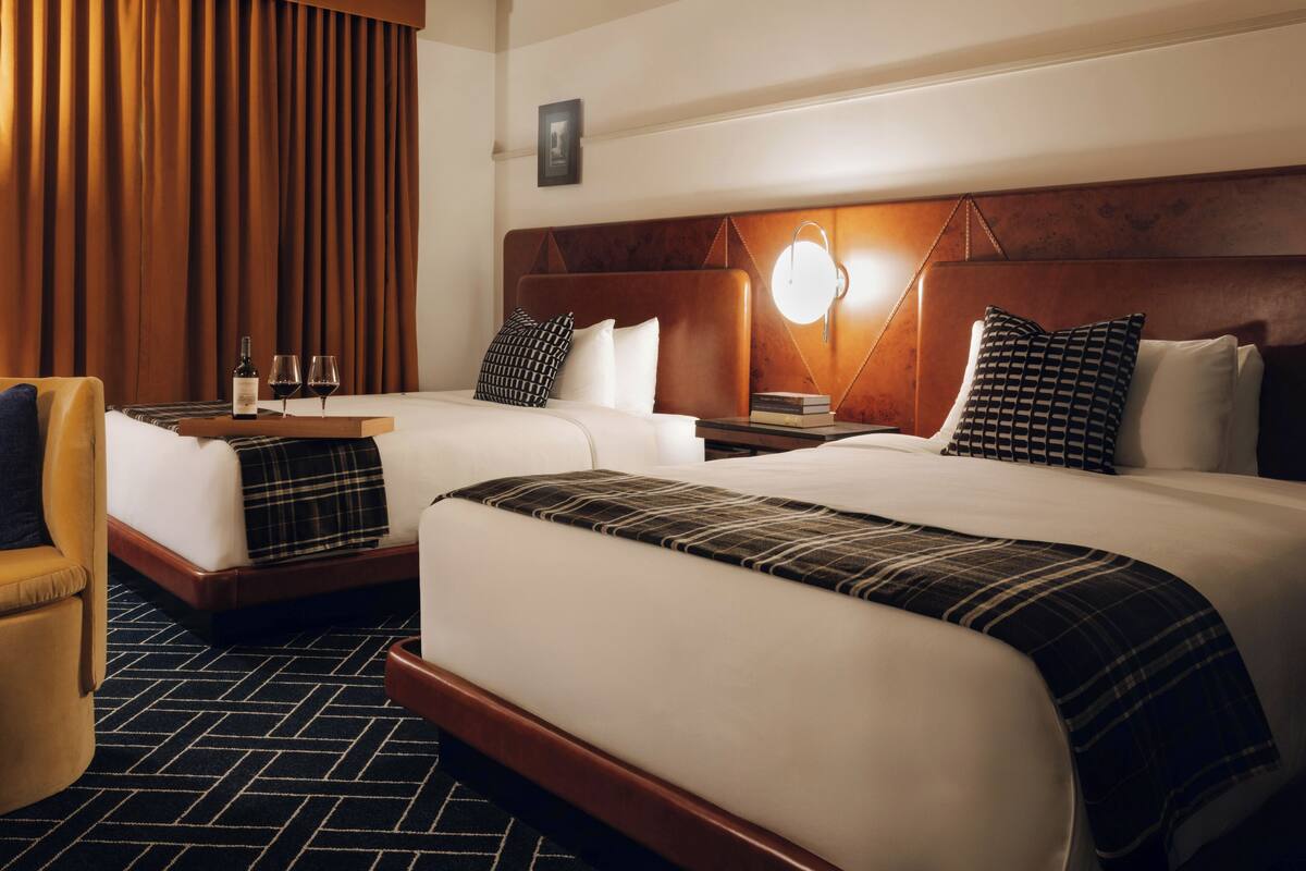 A hotel room with two beds, a wooden headboard, a nightstand, a lamp, a chair, and a large window with curtains.