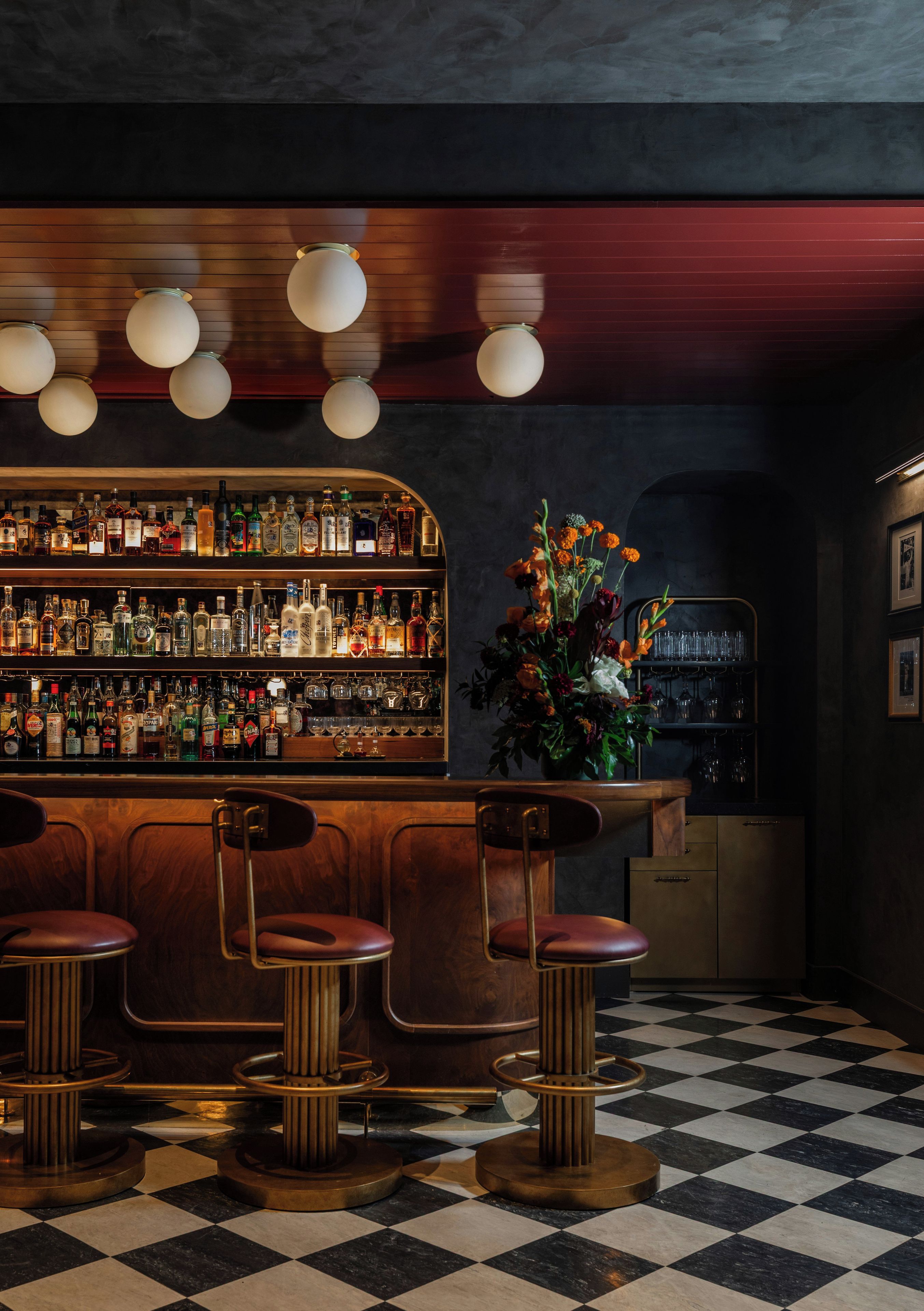 Un bar con piso a cuadros, taburetes de bar, una estantería con botellas de licores y un arreglo floral.
