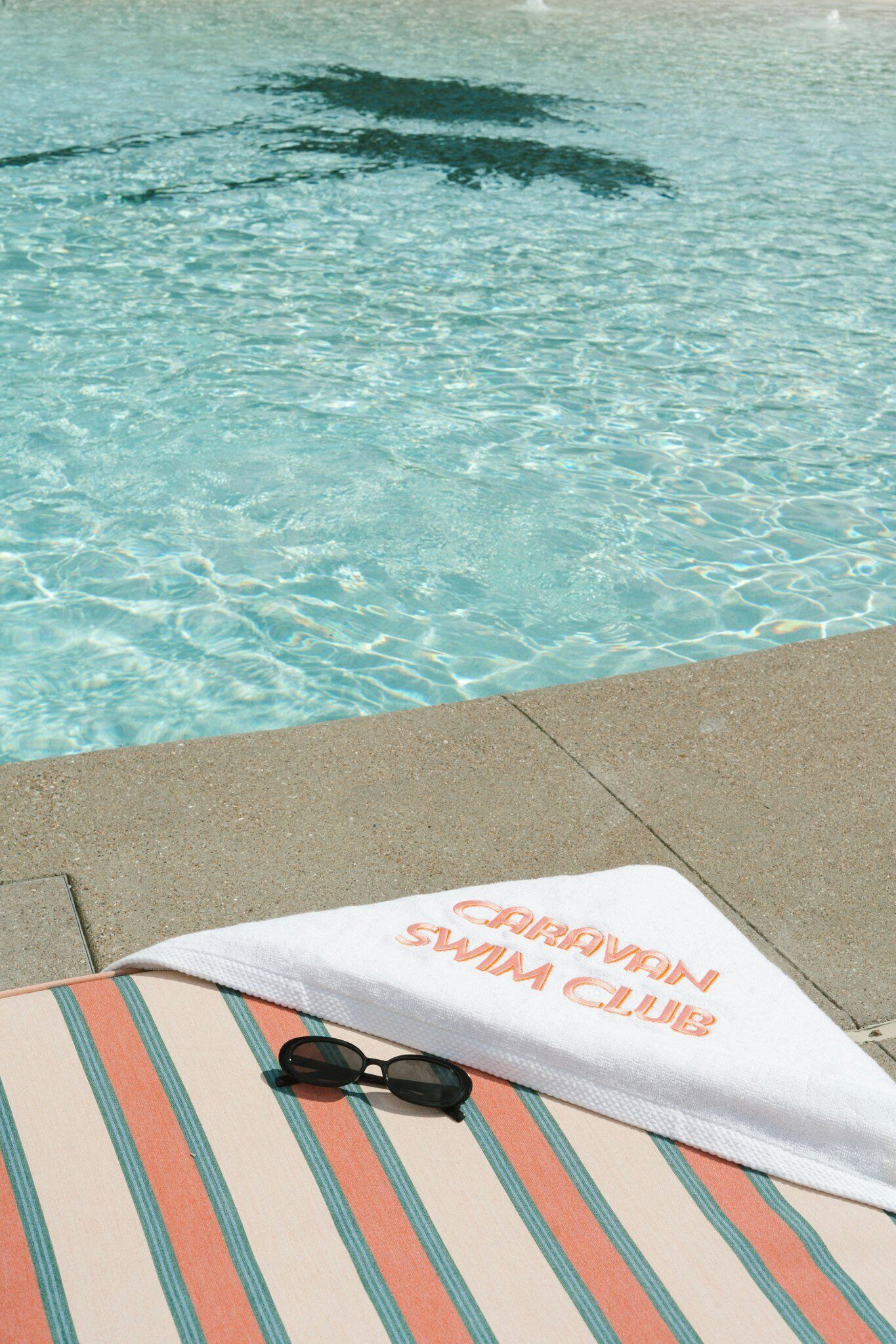A poolside area with a striped towel and sunglasses on a lounge chair, and a towel with 'CARAVAN SWIM CLUB' text.