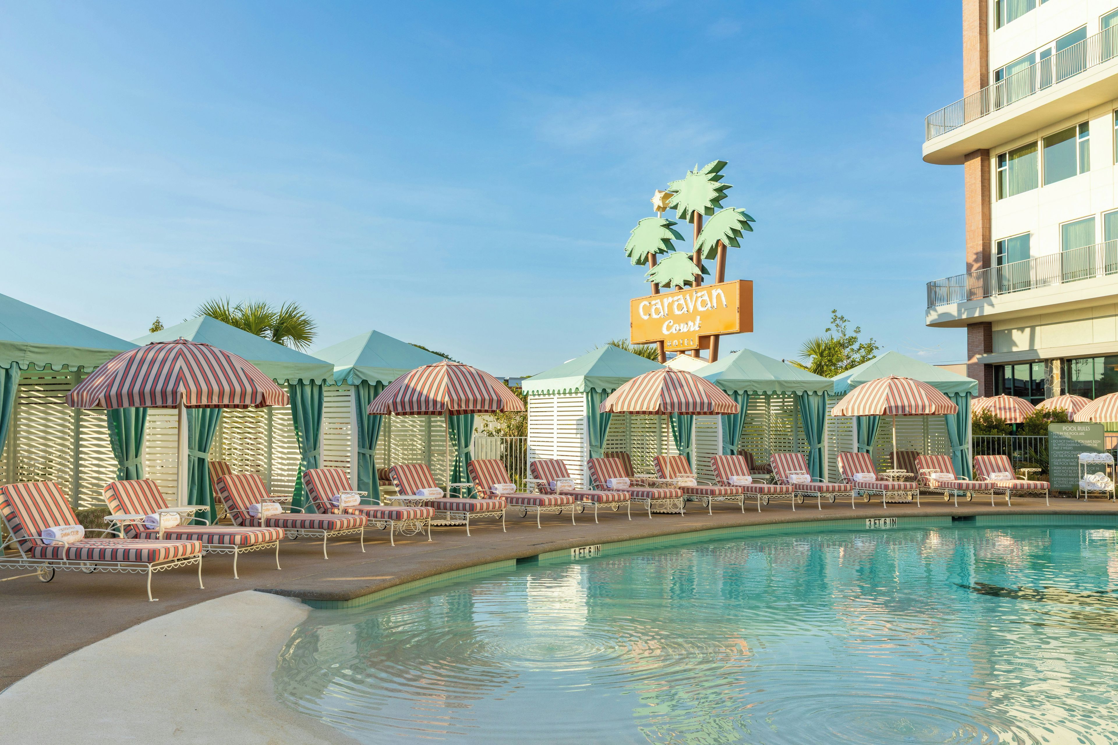 Un área de piscina con sombrillas a rayas y sillones de descanso, un cartel del Hotel Caravan Court y un cielo azul despejado.