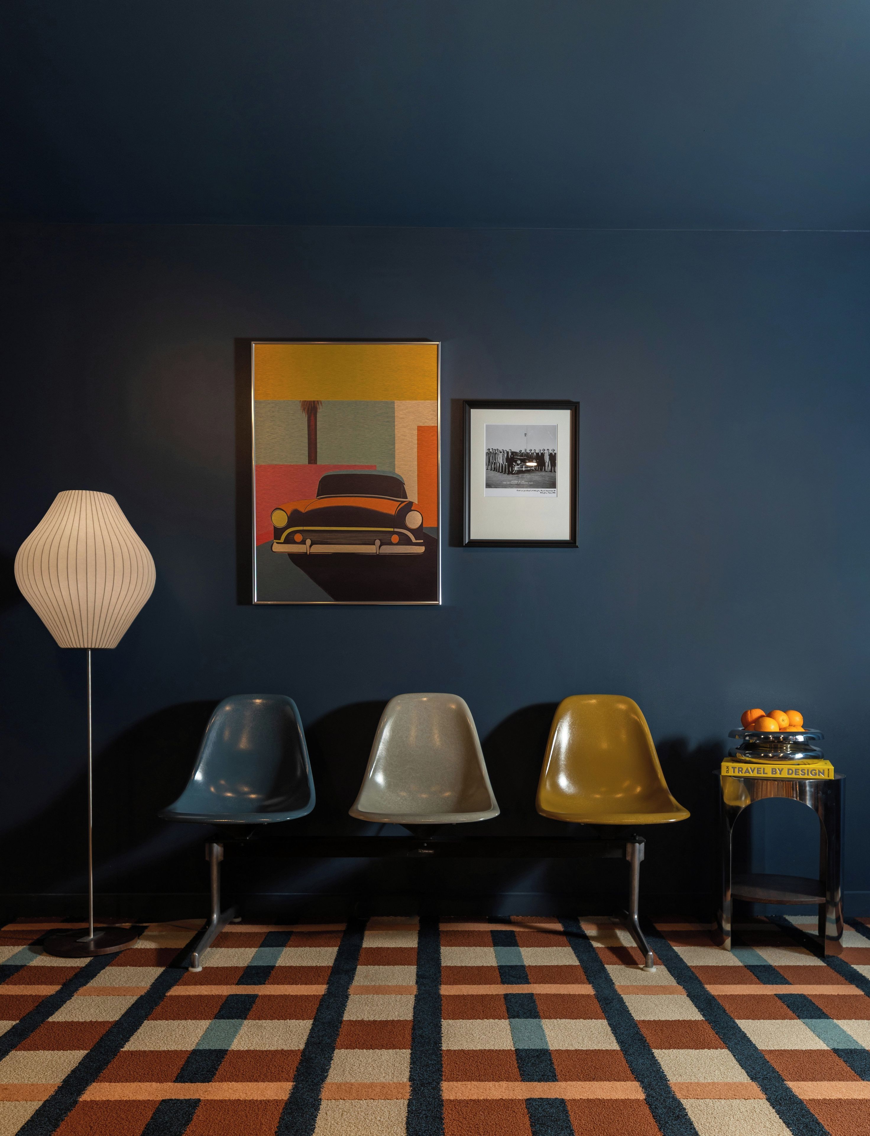 A room with a dark blue wall, a patterned carpet, three chairs, a lamp, and two framed pictures.