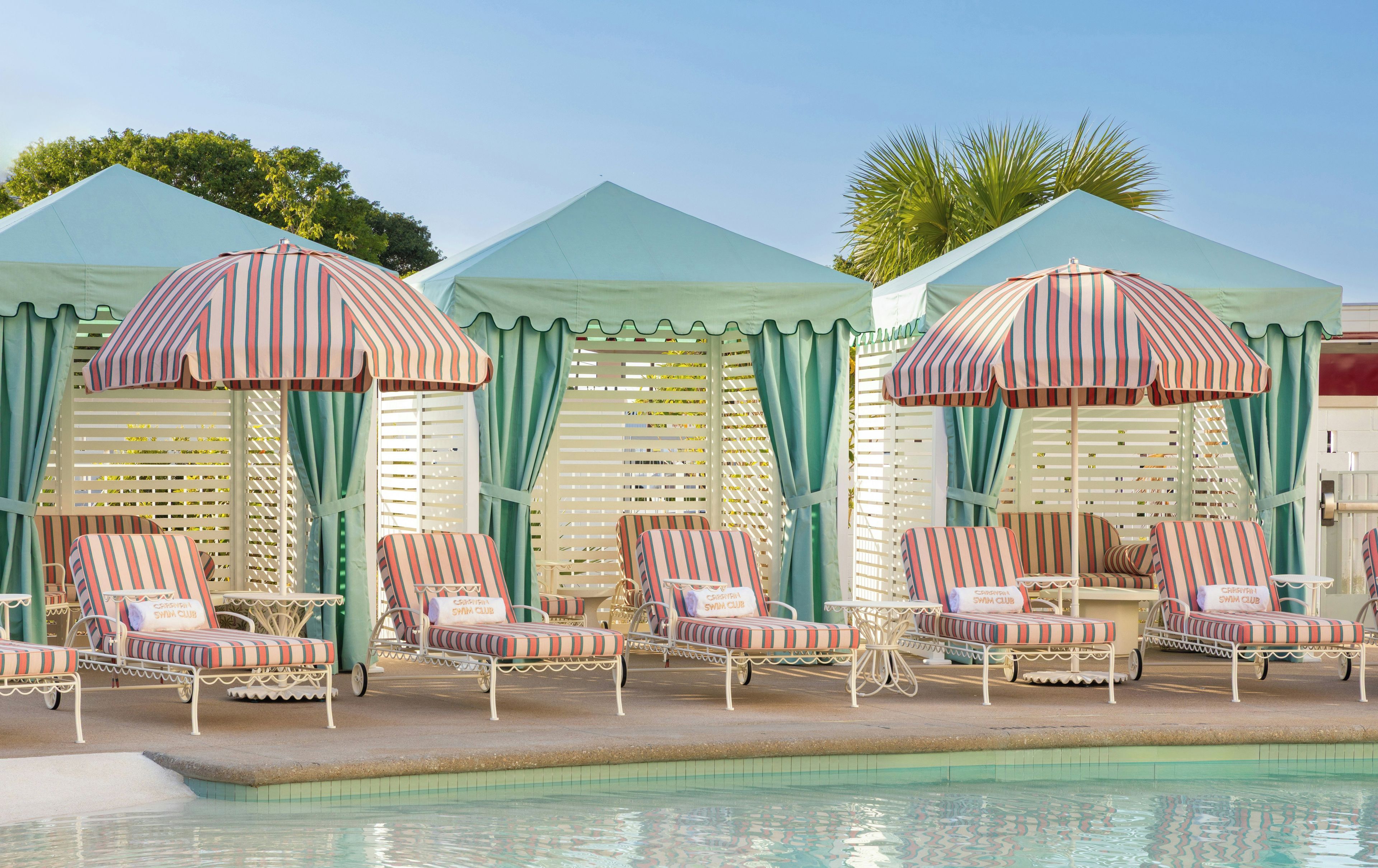 A poolside area with striped lounge chairs and umbrellas, a tiled pool, and a building with a peaked roof.