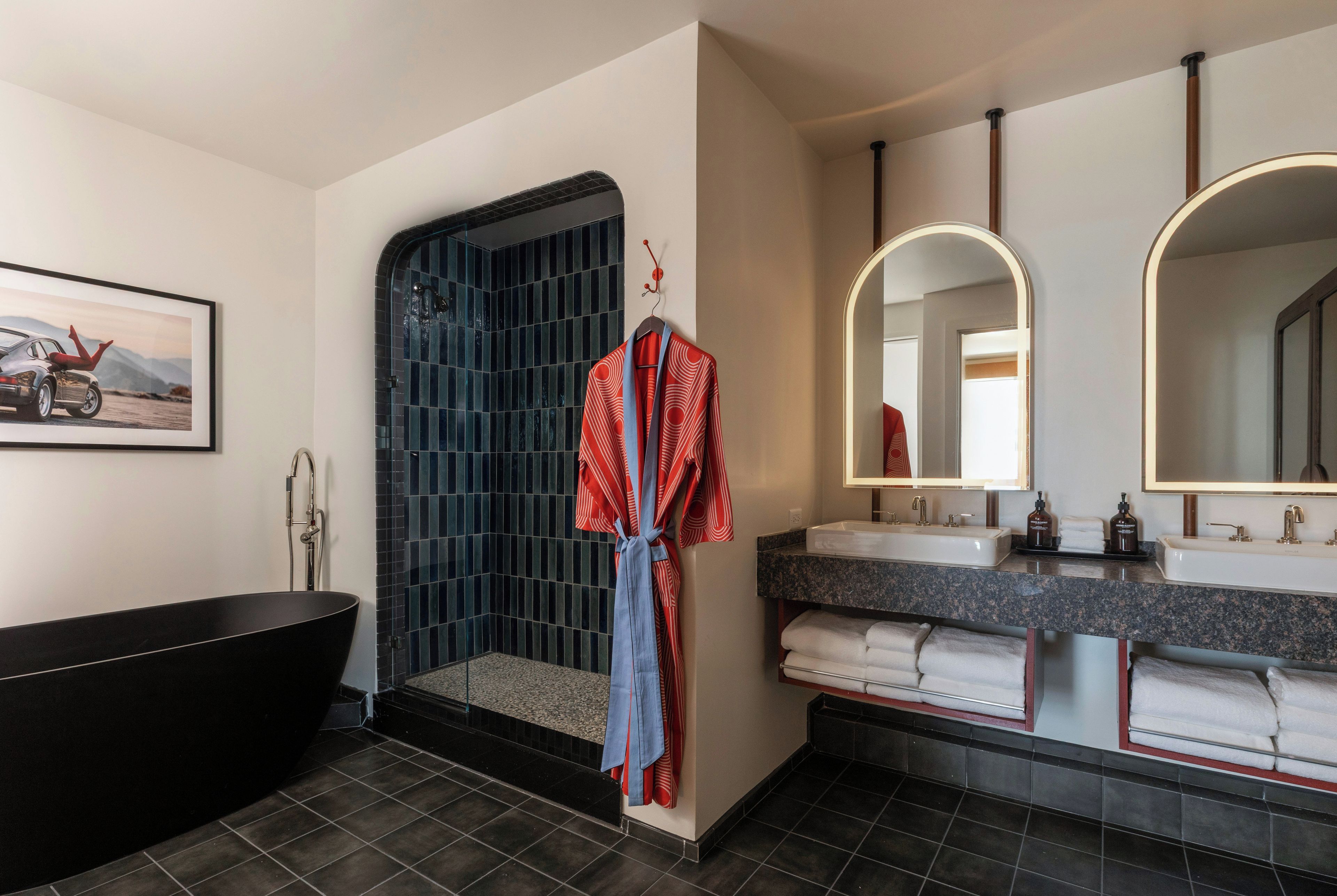 A modern bathroom with a black bathtub, a walk-in shower with blue tiles, a large mirror, and a framed picture on the wall.