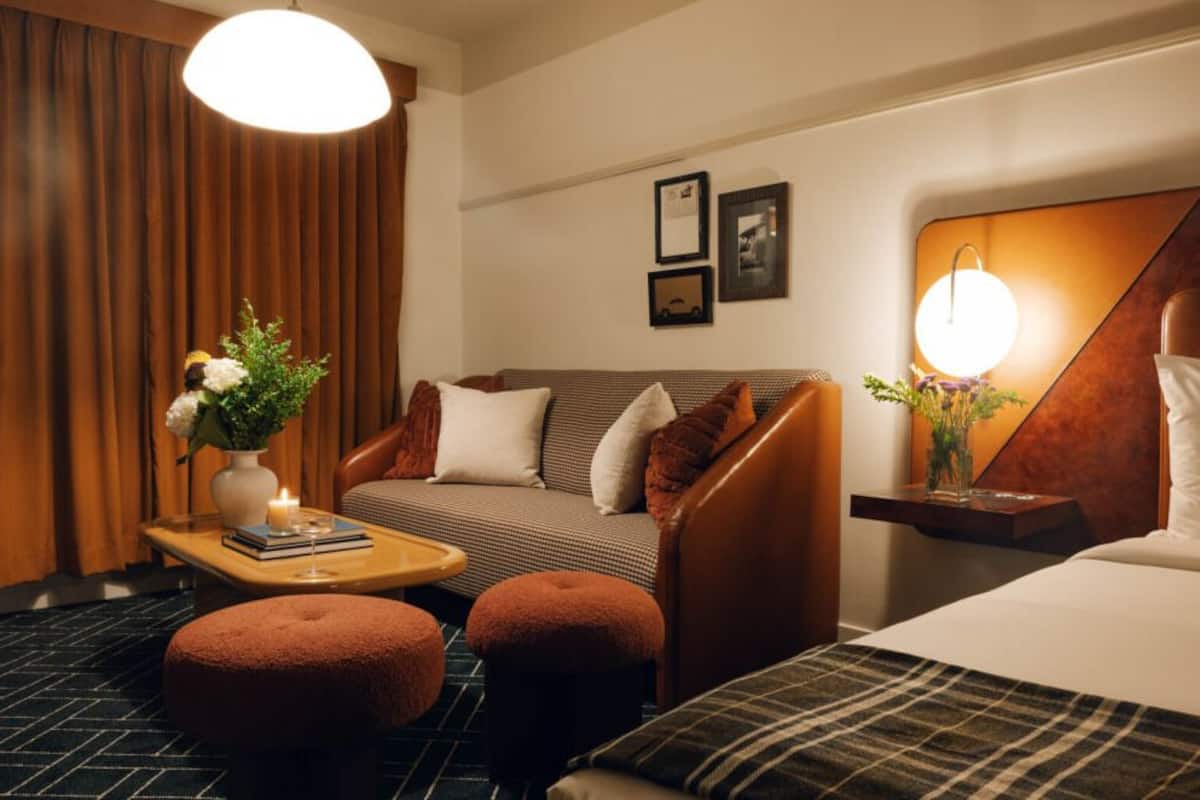 A hotel room with a sofa, ottomans, a bedside table with a vase of flowers, a lamp, and framed pictures on the wall.