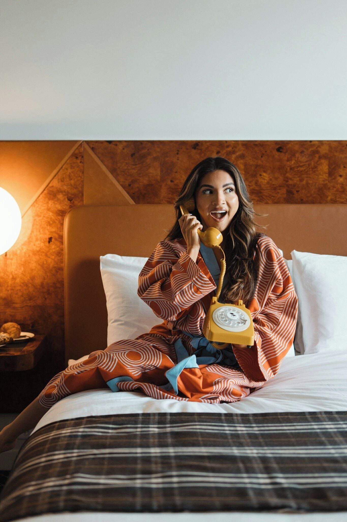 A person sitting on a bed with a telephone, a pillow, and a bedside table with a lamp.