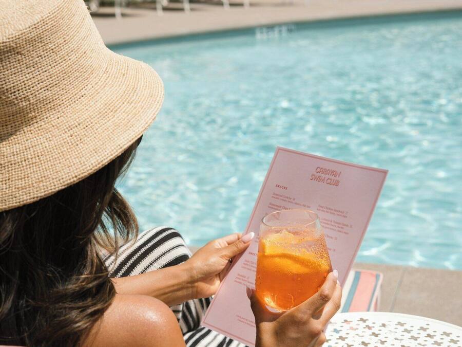 A person sitting by a poolside table with a menu, holding a drink.
