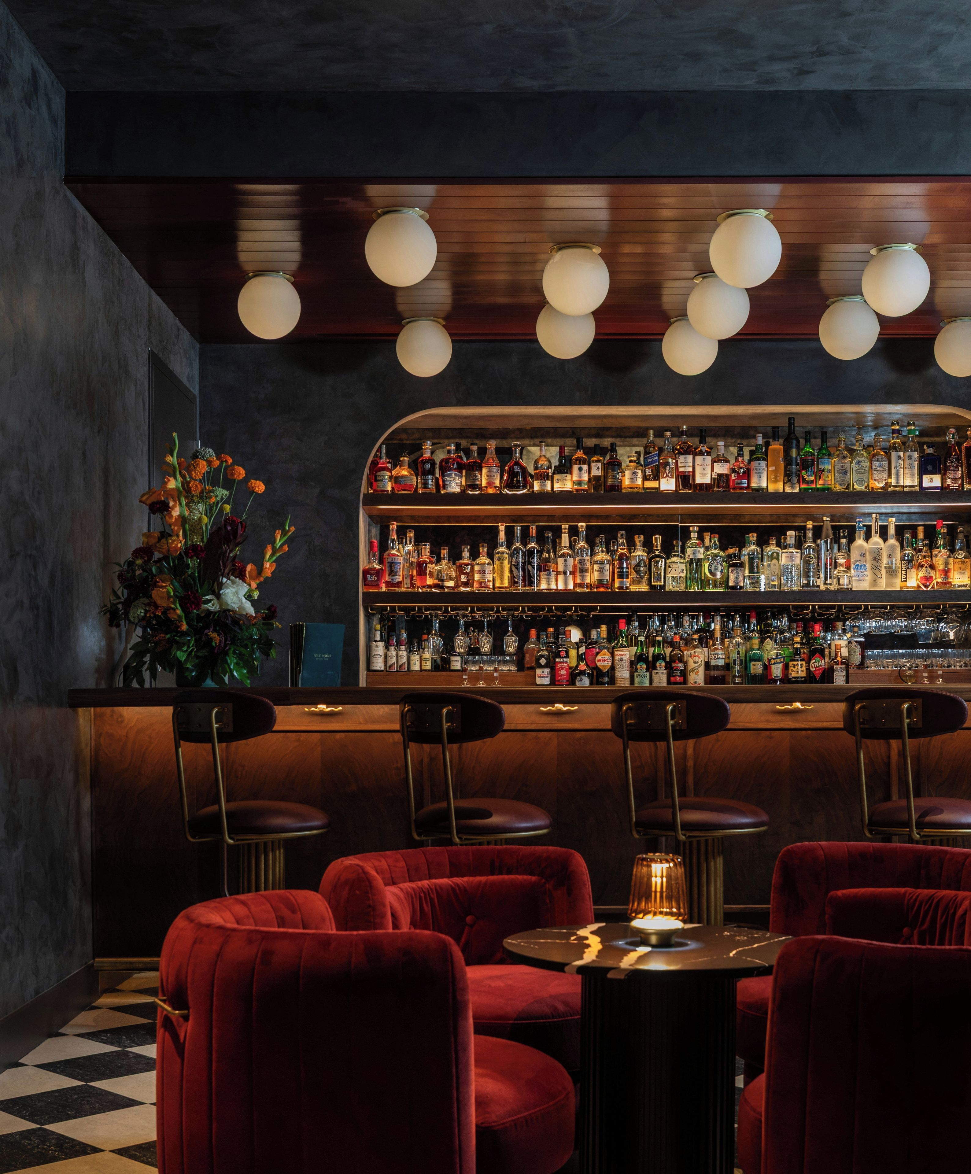 A bar area with multiple shelves of liquor bottles, red upholstered chairs, and a decorative plant.