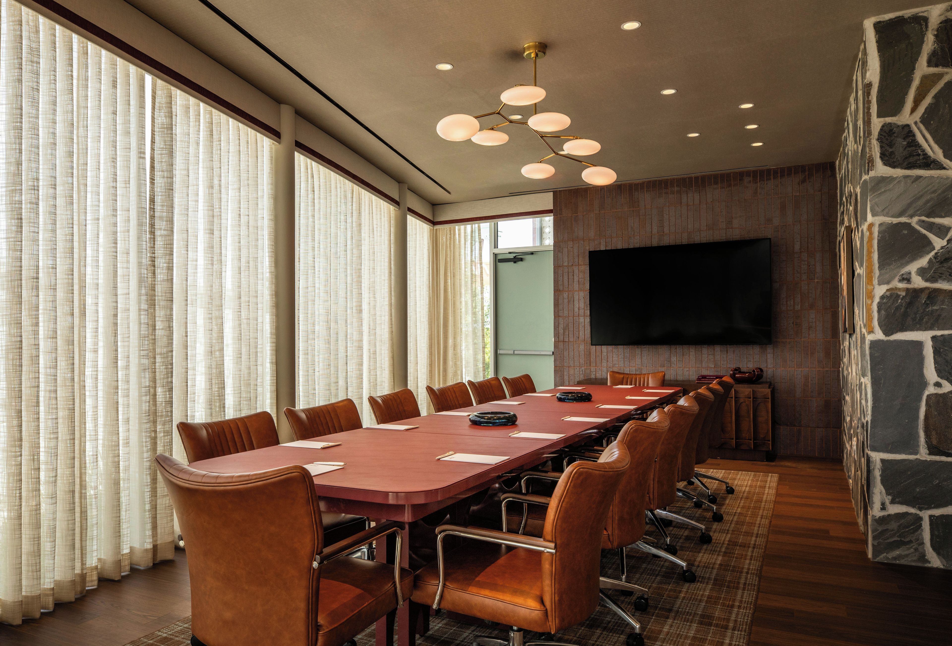 A conference room with a long table, leather chairs, a flat-screen TV on the wall, and large windows with curtains.