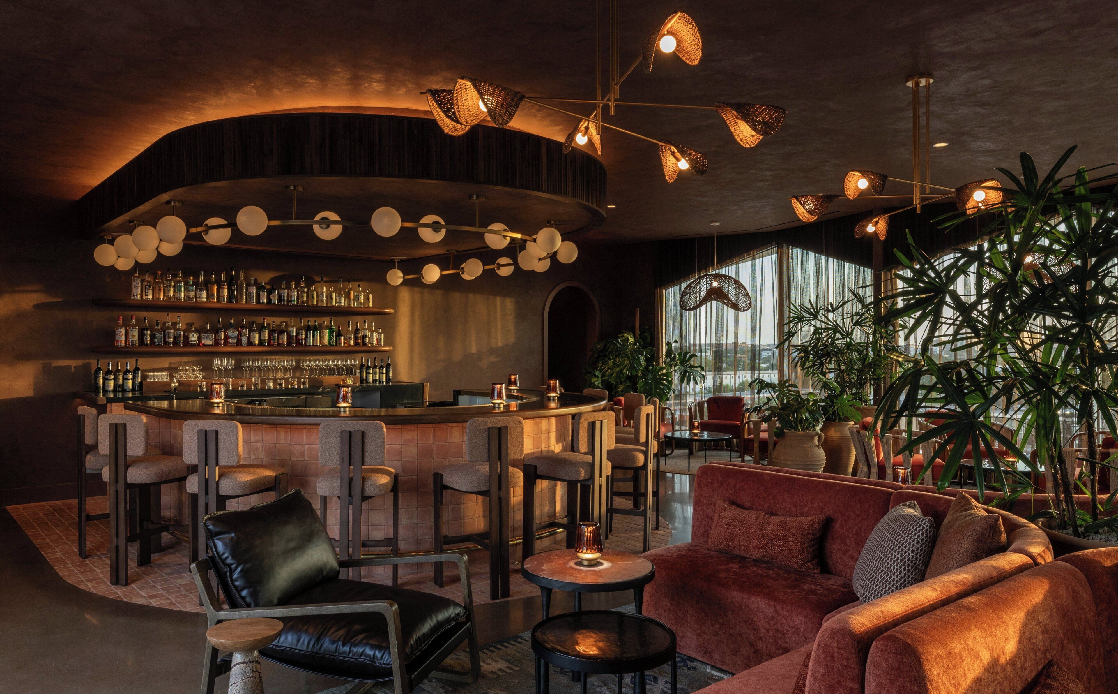 A bar area with a curved counter, multiple seating options including leather chairs and sofas, and a variety of plants.