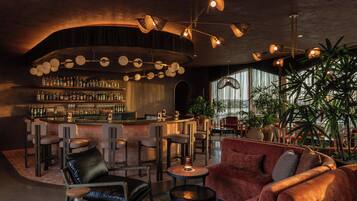 Zona de bar con una barra curva, diversas opciones de asientos, incluyendo sillones y sofás de cuero, y una variedad de plantas.