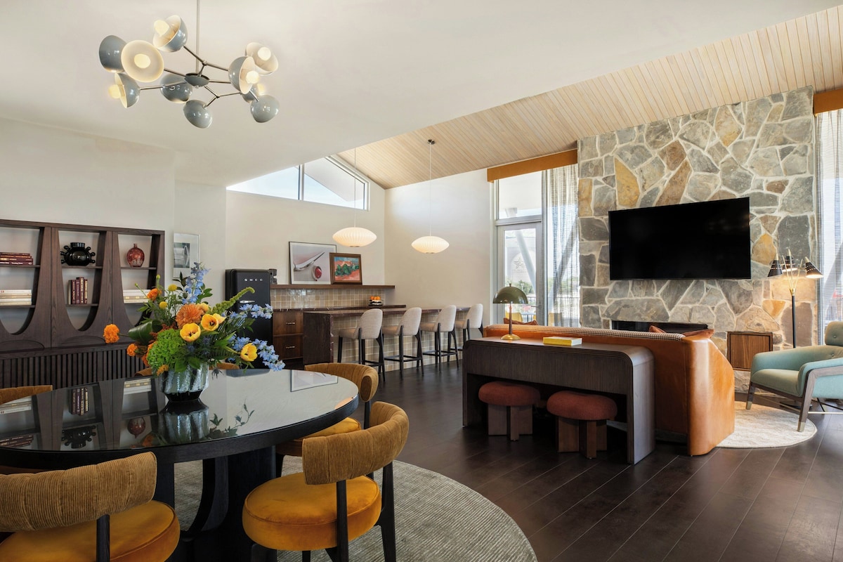A modern living room with a stone fireplace, a dining area with a round table, and a kitchen area.