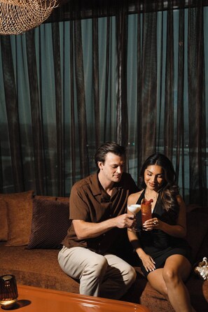 A couple sitting on a couch with a drink in hand, a candle on a table, and a woven light fixture above. - Caravan Court Hotel (Arlington)