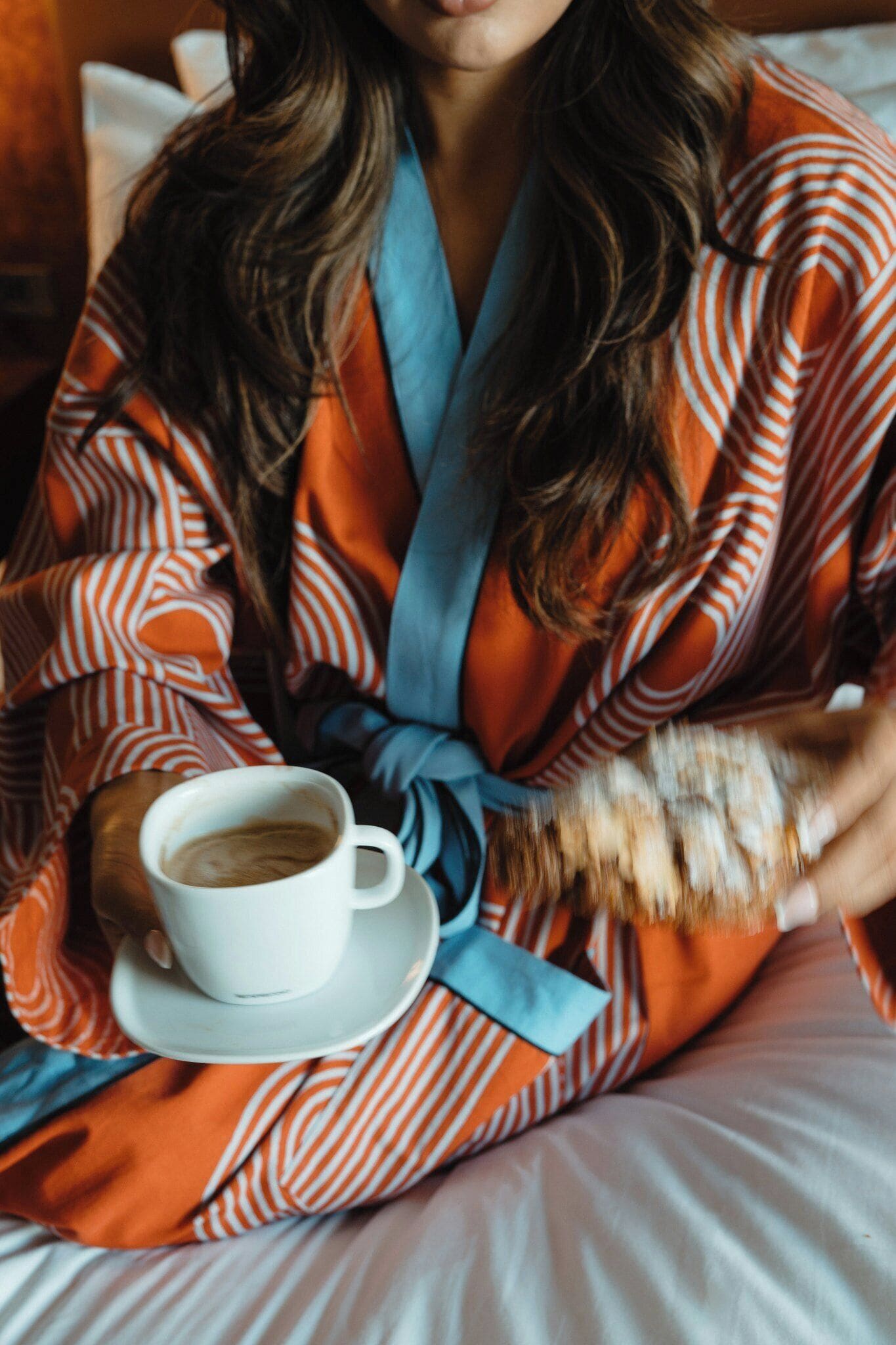 A person in a striped robe holding a cup of coffee on a bed.