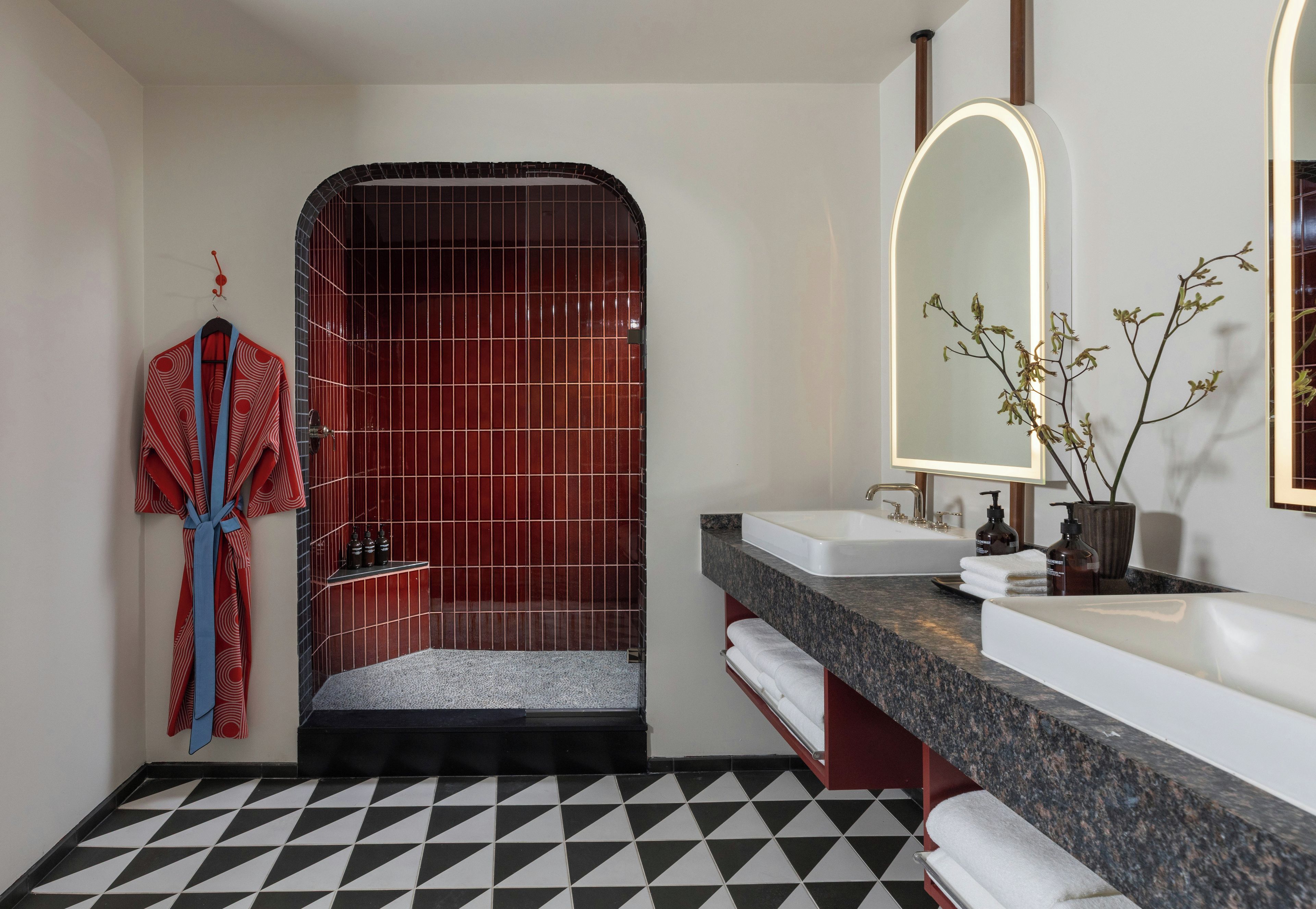 A modern bathroom with a checkered floor, a shower with a red-tiled wall, a vanity with a marble countertop, and a mirror with a light.