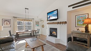 Een woonkamer met een open haard, een televisie erboven en een eethoek met een tafel en stoelen.