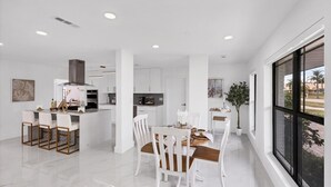 Una cocina moderna con armarios blancos, una isla central y una zona de comedor con sillas blancas y una mesa de madera.