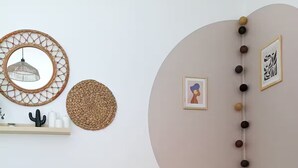 Ein Zimmer mit einem runden Holztisch, einem weißen Stuhl mit Kissen, einem wandmontierten Spiegel und Dekorationsobjekten, darunter ein gewebter Korb und eine Perlenkette.