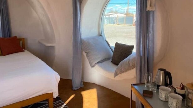 Ein gemütliches Zimmer mit einem Bett, einer Sitzgelegenheit am Fenster mit Kissen und einem Tisch mit Wasserkocher und Tassen.