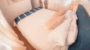 Quarto de mobil home compacto, com cama de solteiro, almofadas, cortinas e um quadro na parede.