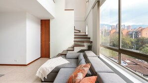 A modern living room with a grey sectional sofa, a wooden staircase, and a large window offering a city view. - Apartamentos SETAI (Medellín)