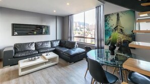 A modern living room with a black leather sofa, a glass dining table, and a cityscape painting. - Apartamentos SETAI (Medellín)