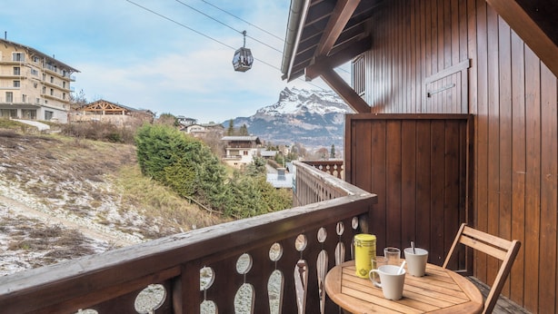 Ein Holzbalkon mit Tisch und Stühlen, mit Blick auf eine Berglandschaft, im Hintergrund ist eine Seilbahn zu sehen.