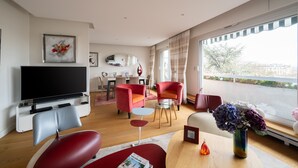 A modern living room with a flat-screen TV, red and grey chairs, a glass coffee table, and a view of the city through large windows. - Appartement Trocadero - 4 étoiles - Welkeys (Paris)