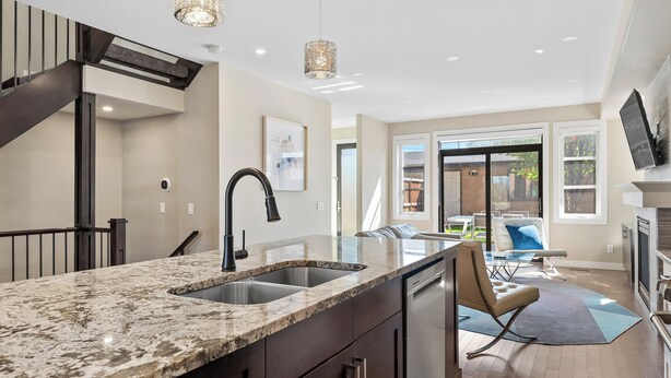 A modern kitchen with a granite countertop, double sink, and stainless steel appliances. There is a dining area with a table and chairs, and a large window providing natural light. - Spacious Central 4BR Duplex with 2 Car Garage & Downtown View from Rooftop Patio (Calgary)