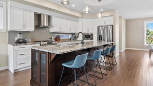 A modern kitchen with white cabinets, stainless steel appliances, a granite countertop, and blue bar stools. - Spacious Central 4BR Duplex with 2 Car Garage & Downtown View from Rooftop Patio (Calgary)