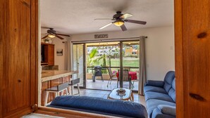 Ein Zimmer mit Deckenventilator, einer Schiebetür zu einer Terrasse, einem blauen Sofa, einem kleinen Tisch und einem Holzschrank.