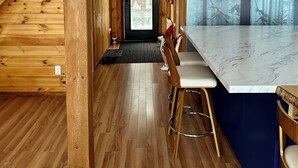 Interior de madera con área de cocina, una escalera y vista a un bosque por una ventana.