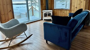 Un acogedor interior de cabaña con una mecedora, un sofá azul, una chimenea blanca y un amplio ventanal con vistas a un paisaje nevado.