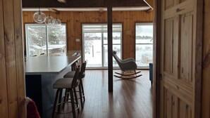 Un interior de madera con una zona de cocina, taburetes de bar, una mecedora y ventanas grandes.