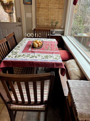 A dining table with a floral tablecloth, wooden chairs, a fruit basket, and a window with a view of trees.