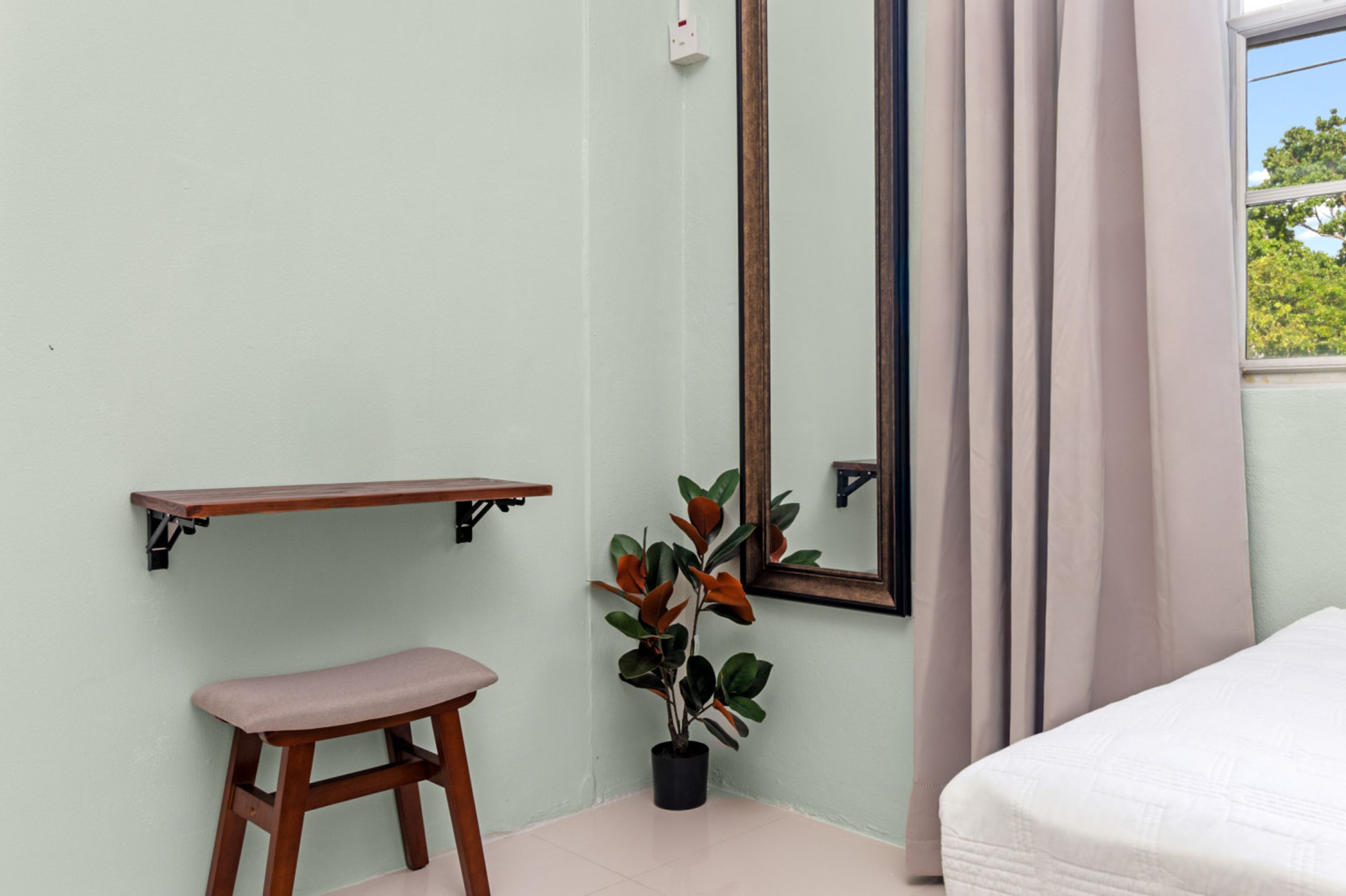 Ein Zimmer mit einem Bett, einem kleinen Hocker, einer Topfpflanze, einem Spiegel und einem Regal.