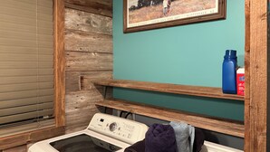 A laundry area with a washing machine, a drying rack, a framed picture, and a shelf with a water bottle and a box. - Cedar Ridge Ranch Barndominium/Cozy Rustic 3-Bedroom with Wood-Burning Stove (Hutchinson)