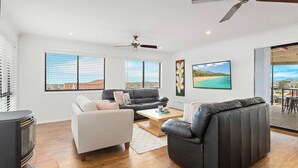 A modern living room with a sofa, armchair, and a coffee table. There is a ceiling fan, a fireplace, and a large window with blinds. - Pacific View - Pet Friendly (Flinders)