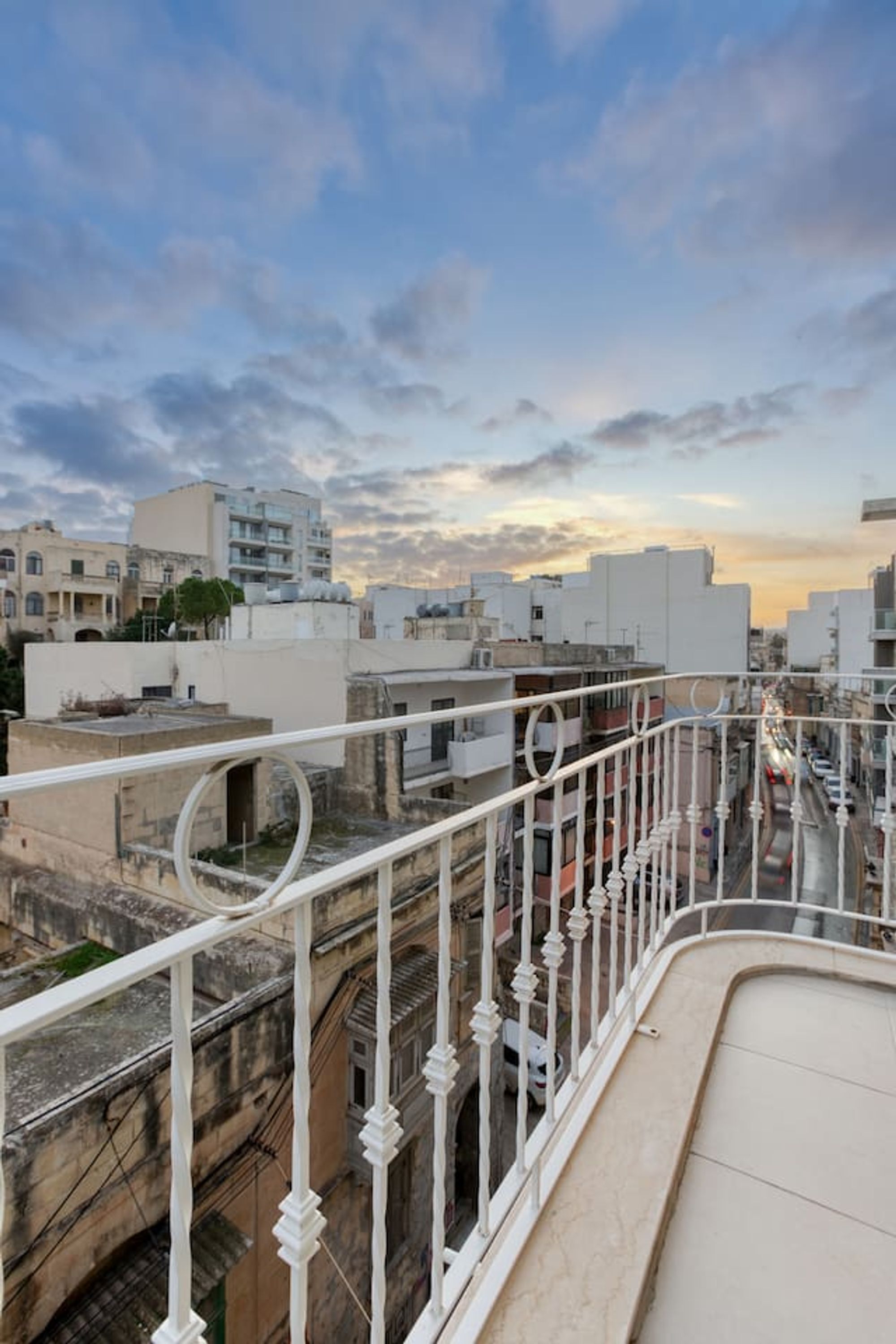 A rooftop terrace with a white balustrade, offering views of surrounding buildings and a sunset sky.