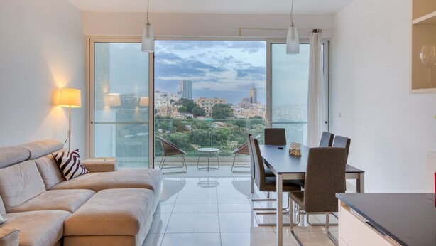 A modern living room with a sofa, dining table, chairs, and a view of a cityscape through large windows. - Valley Sea Views With Furnished BBQ Balcony (St. Julian's)